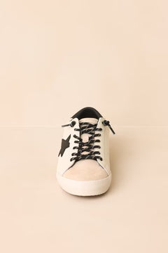 A single low-top sneaker shown from the front on a plain beige background. The shoe is primarily off-white with black laces, a black star detail on the side, and a black collar lining.