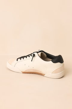 A low-top sneaker in off-white leather with black laces, a black heel tab, and a light brown gum sole, shown from a side profile on a warm beige background.