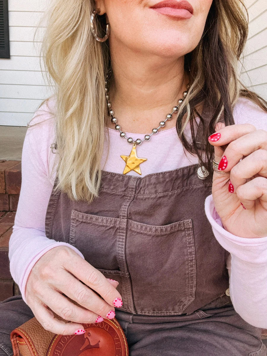 Person wearing a star-shaped necklace on a pink shirt with brown overalls.