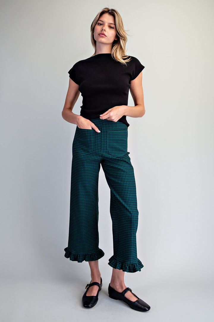 A woman modeling dark green and black checkered cropped pants with ruffled hems, paired with a black short-sleeve top and black Mary Jane shoes, against a light gray studio background.
