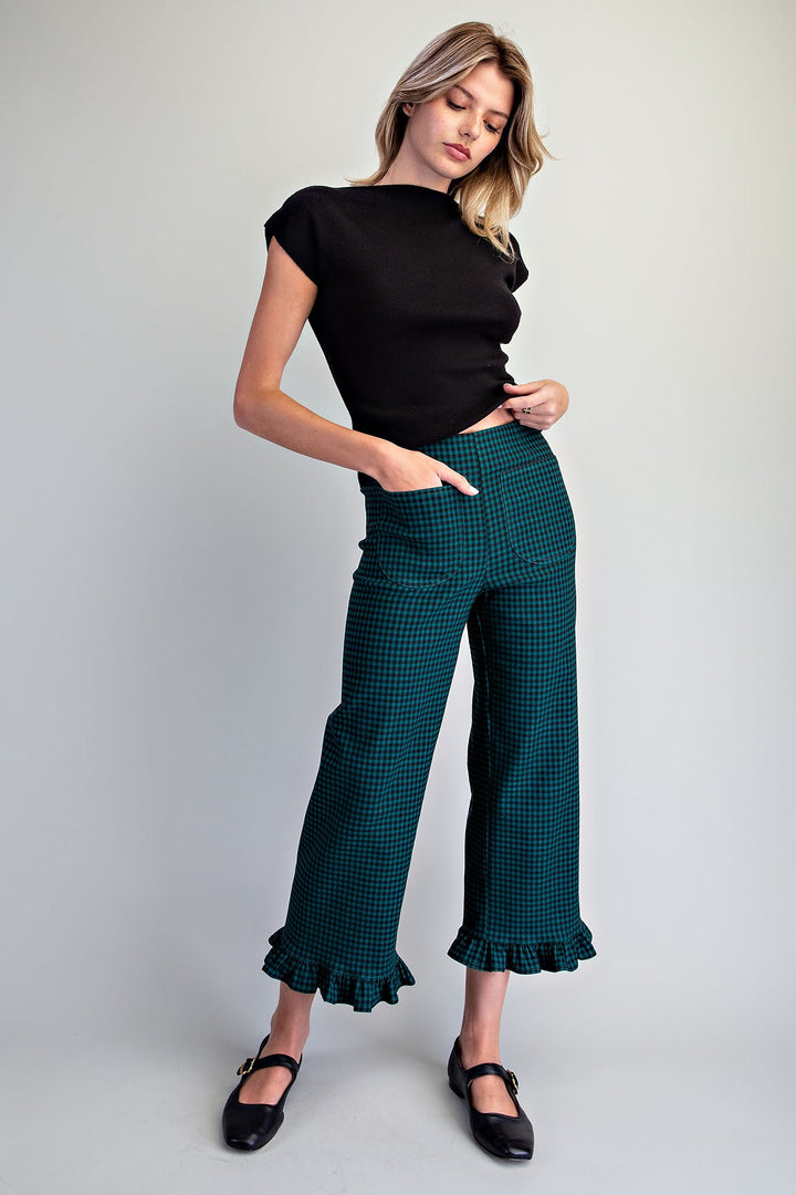 A woman modeling a black short-sleeve top and dark green and black checkered cropped pants with ruffled hems, paired with black Mary Jane shoes, against a light gray studio background.
