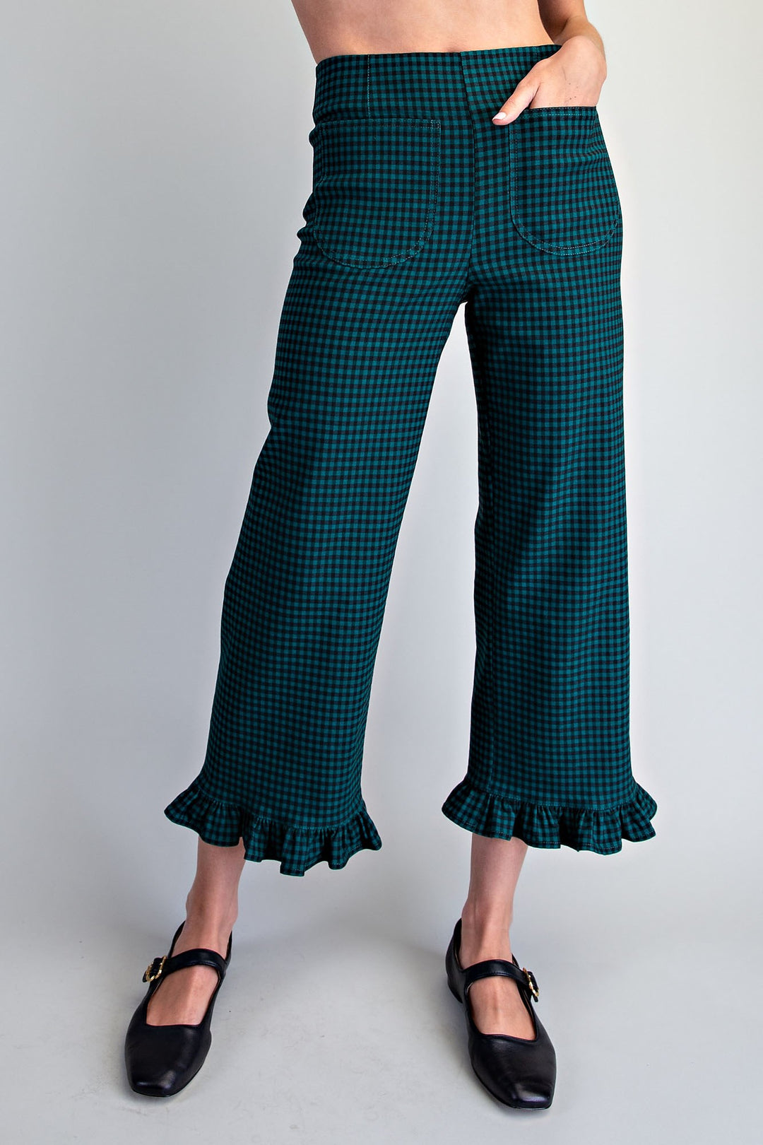 A model is wearing dark green and black checkered cropped pants with a ruffled hem, paired with black Mary Jane shoes, against a light gray studio background.