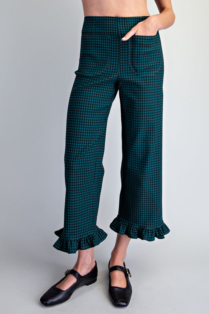 A model is shown from the waist down wearing dark green and black checkered cropped pants with ruffled hems, paired with black Mary Jane shoes, against a light gray studio background.