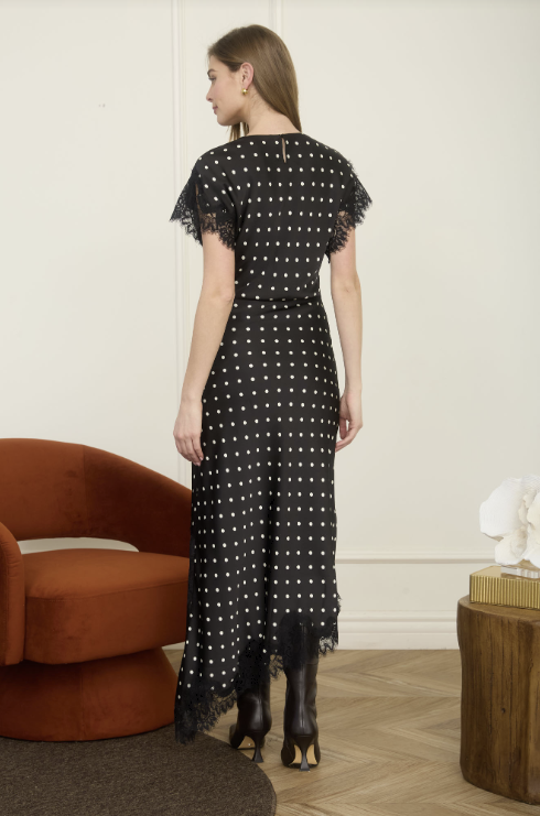 A woman stands with her back to the camera wearing a black midi dress with white polka dots and tattered lace trim on the sleeves and hem, styled with black boots in a modern room setting.