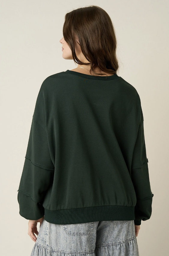 A woman shown from the back wearing a dark forest green cropped sweatshirt with long sleeves and a ribbed hem, paired with light gray tiered pants, against a plain beige background.