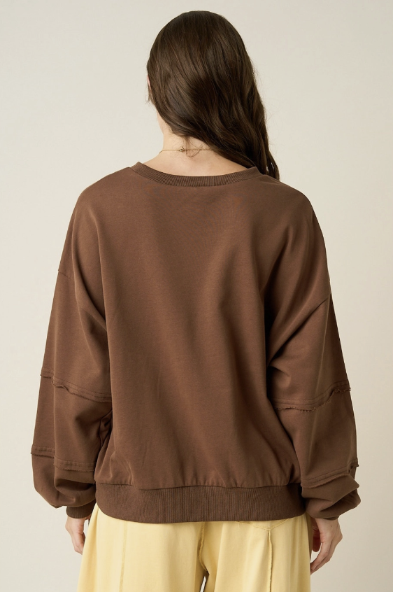 A woman viewed from the back wearing a brown crewneck sweatshirt with long sleeves and light yellow pants, set against a plain light beige background.