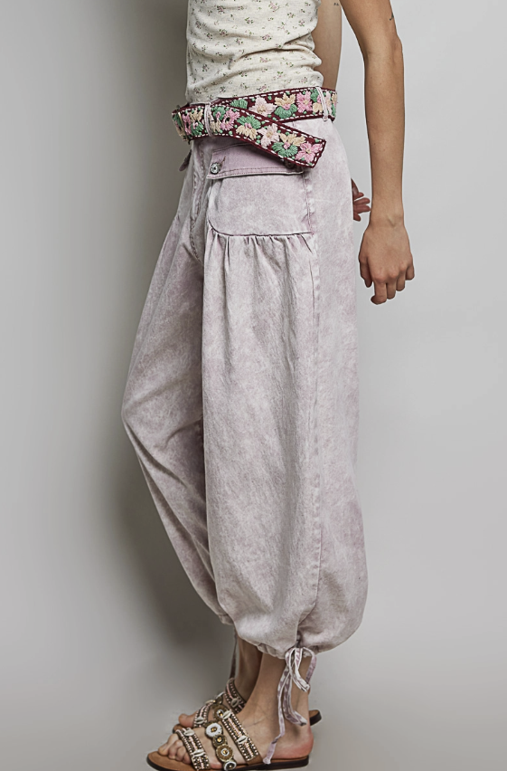 A person wearing loose, baggy light gray harem-style pants with a wide floral waist belt, paired with a cream top and brown sandals, photographed against a plain light gray studio background.