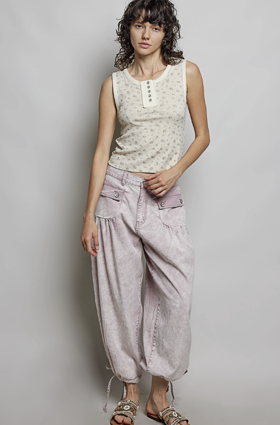 A model stands against a light gray studio background wearing a sleeveless cream top with a small floral print and a button placket, paired with loose light pink baggy pants and metallic sandals.