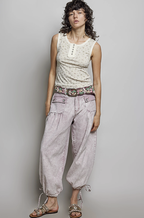 A model stands against a light gray studio background wearing a sleeveless cream top with a small floral print, loose light pinkish-purple baggy pants, a patterned belt, and brown sandals.
