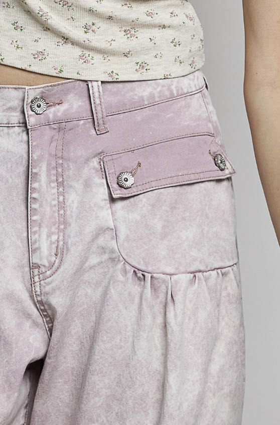 Close-up of a person wearing light pink washed denim pants with a decorative front pocket and silver-tone button details, paired with a cream floral top.