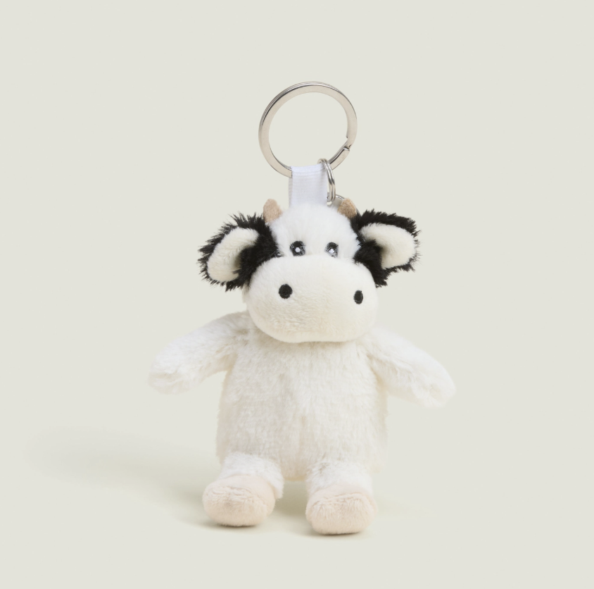 A small plush cow keychain with a white body, black patches on the ears and face, and a silver metal key ring, photographed on a plain light beige background.