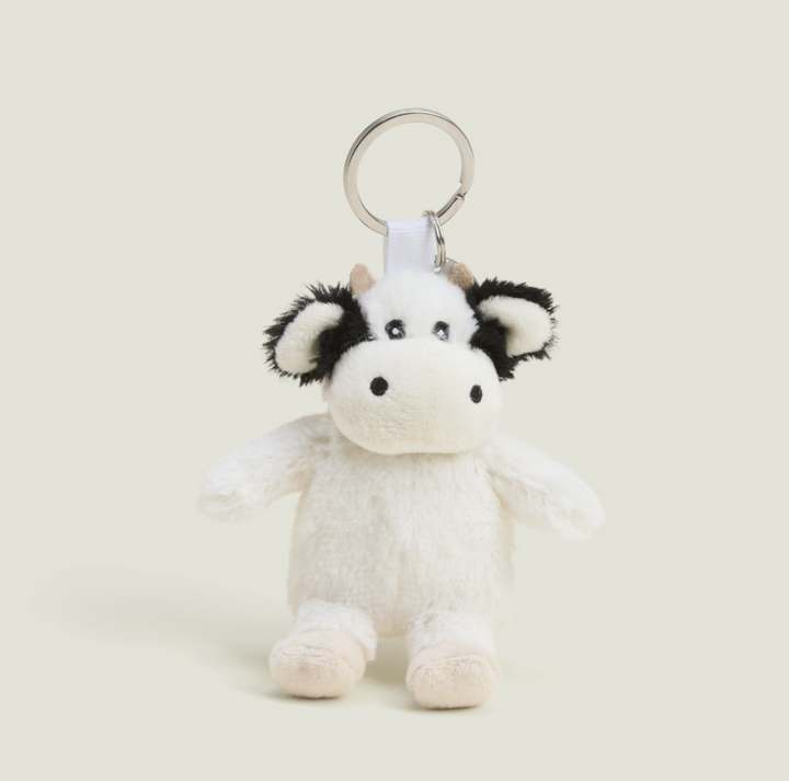 A small plush cow keychain with a white body, black patches on the ears and face, and a silver metal key ring, photographed on a plain light beige background.