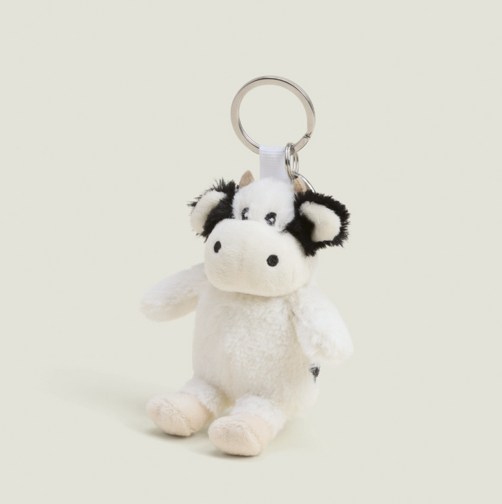 A small plush cow keychain with a white body, black patches on the ears and eyes, and a silver metal key ring, photographed on a plain light beige background.