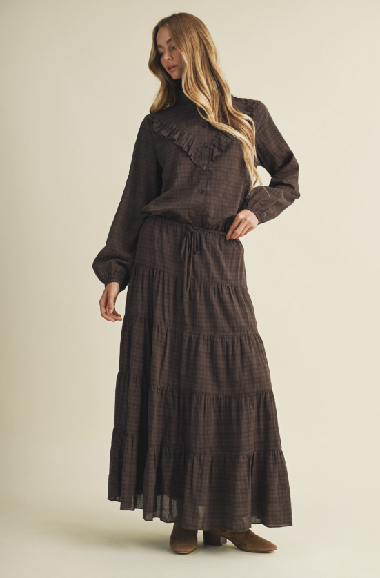 A woman modeling a dark brown plaid long-sleeve blouse and matching tiered maxi skirt set, paired with brown boots, against a solid light beige background.