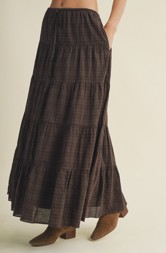 A model wearing a dark brown tiered maxi skirt with a subtle plaid pattern and brown ankle boots, shown from the waist down against a solid light beige background.