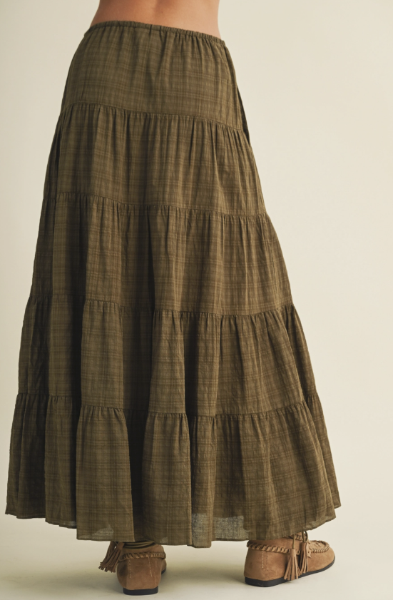An olive green tiered maxi skirt with a subtle plaid pattern, shown on a model from the back wearing brown suede moccasin-style boots against a light beige studio background.