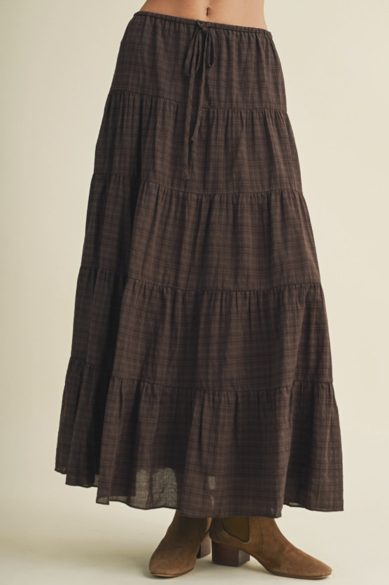 A dark brown tiered maxi skirt with a subtle plaid pattern, featuring a drawstring waist and ruffled hems, shown on a model wearing brown suede boots against a light beige background.