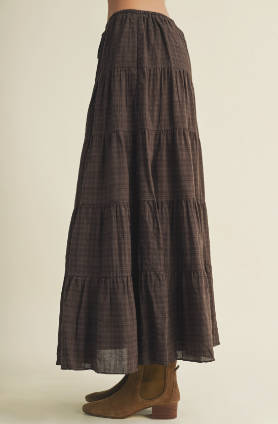 A model is shown from the waist down wearing a dark brown and black tiered maxi skirt with a subtle plaid pattern, paired with brown suede ankle boots, against a solid light beige background.