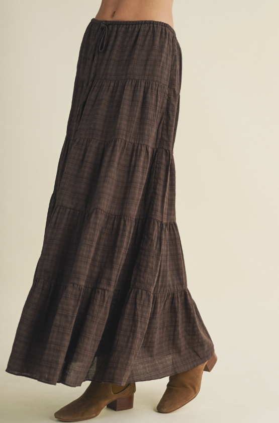 A dark brown tiered maxi skirt with a subtle plaid pattern, shown on a model from the waist down wearing brown suede boots against a light beige background.
