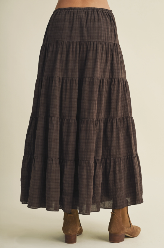 A dark brown tiered maxi skirt with a subtle plaid pattern, shown from the back on a model wearing brown suede ankle boots against a light beige background.