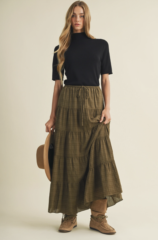 A woman modeling a black short-sleeve mock neck top and an olive green plaid tiered maxi skirt, holding a brown wide-brim hat against a solid light beige background.