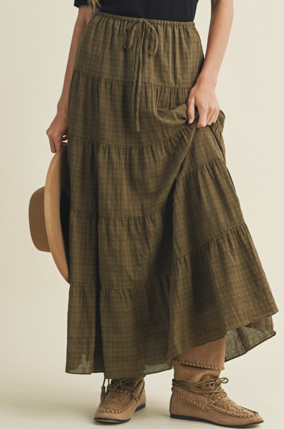 A model wearing a tiered olive green plaid maxi skirt with a drawstring waist, paired with brown lace-up boots and holding a wide-brimmed tan hat against a solid light beige background.