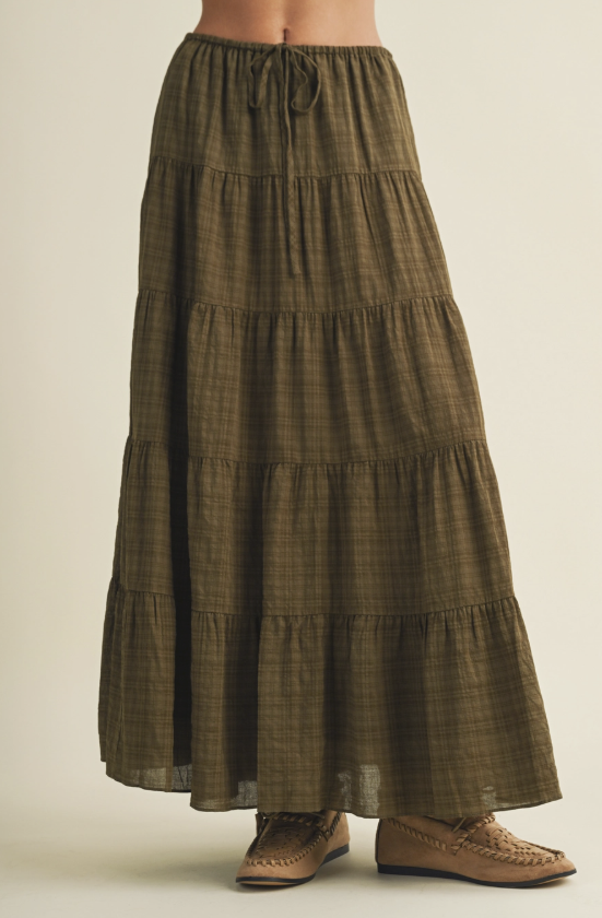 A tiered maxi skirt in an olive green and brown plaid pattern, featuring an elastic drawstring waist and a flowing silhouette, shown on a model wearing brown woven shoes against a light beige background.