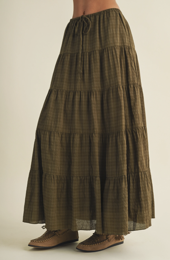 An olive green tiered maxi skirt with a subtle plaid pattern, worn by a model with brown leather moccasin-style shoes against a plain beige studio background.