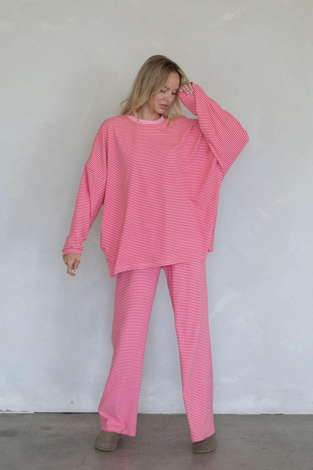 A woman modeling a matching pink and white striped lounge set consisting of a long-sleeve top and wide-leg pants, standing against a light gray textured wall.