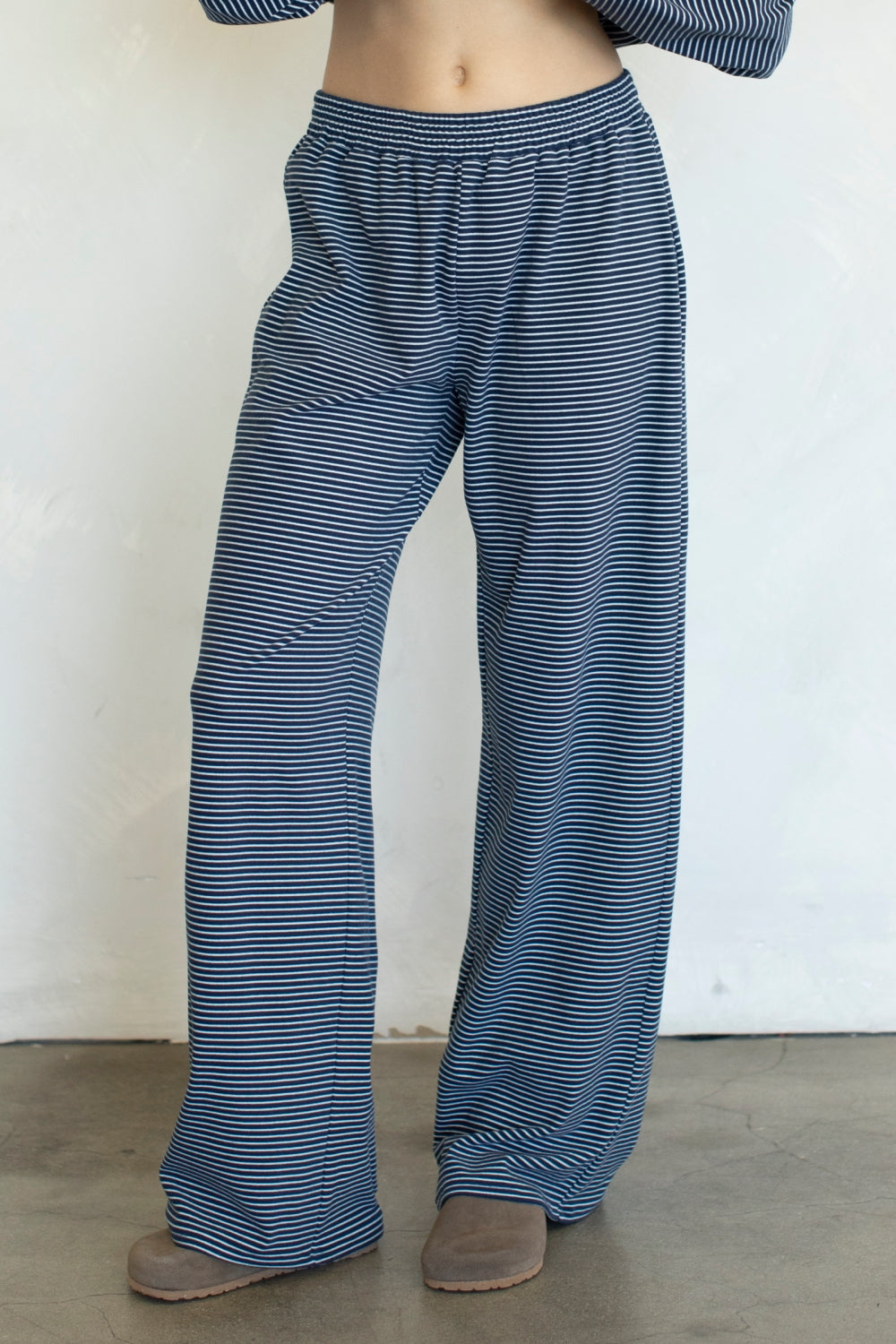 A person wearing loose, wide-leg pants with thin horizontal navy and white stripes, paired with brown slip-on shoes against a plain off-white wall.