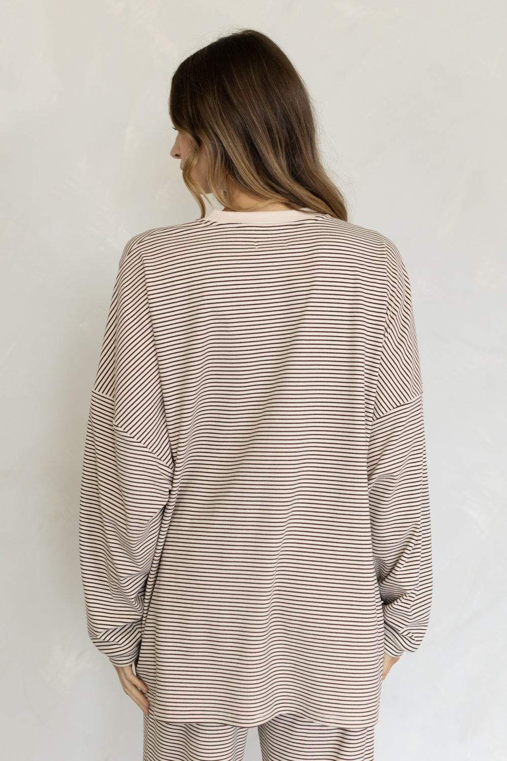 A woman viewed from behind wearing a loose, long-sleeve striped top and matching striped pants in a neutral beige and brown color palette, set against a plain off-white wall.