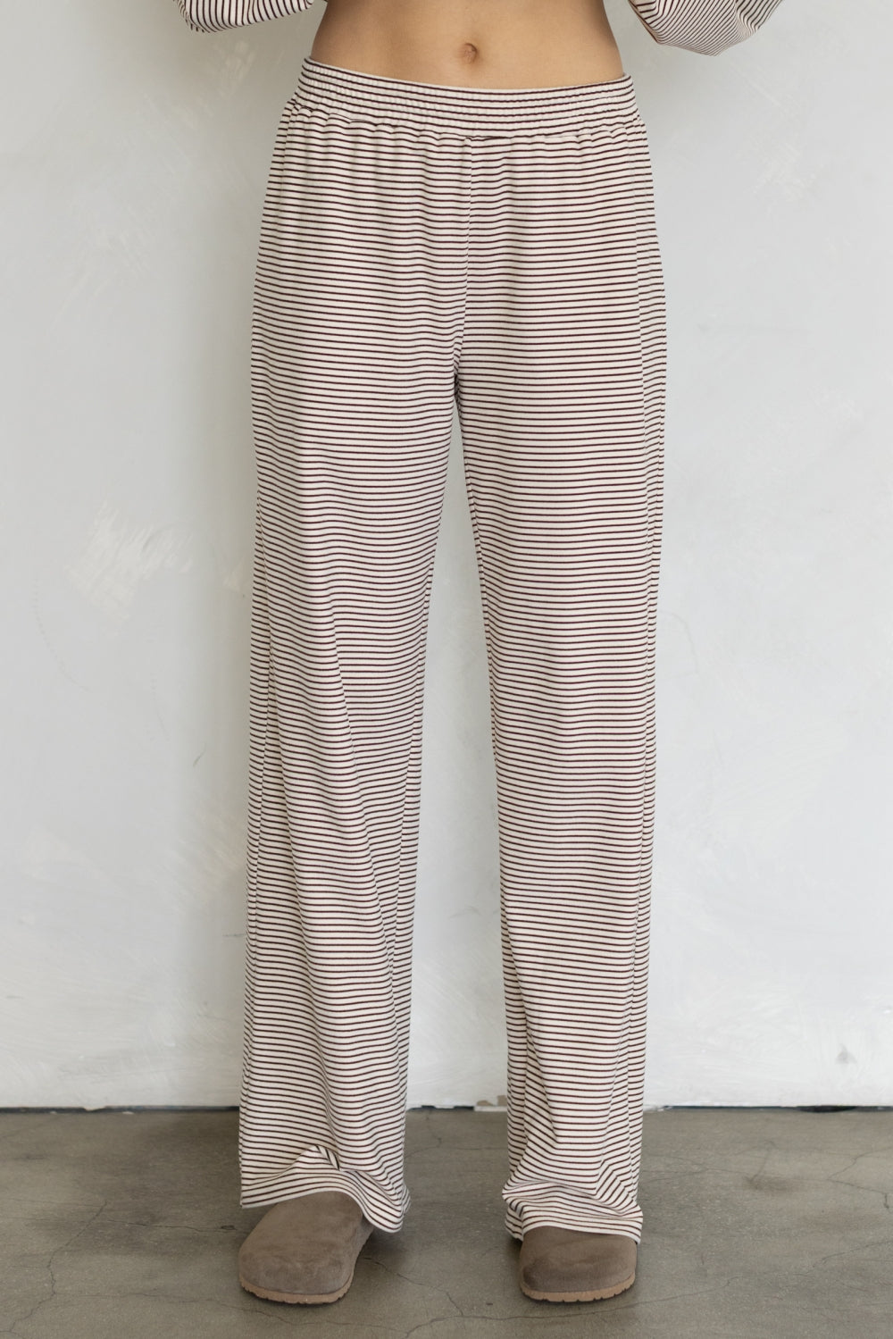 A person wearing loose, wide-leg pants with thin horizontal beige and brown stripes, standing against a plain light wall.