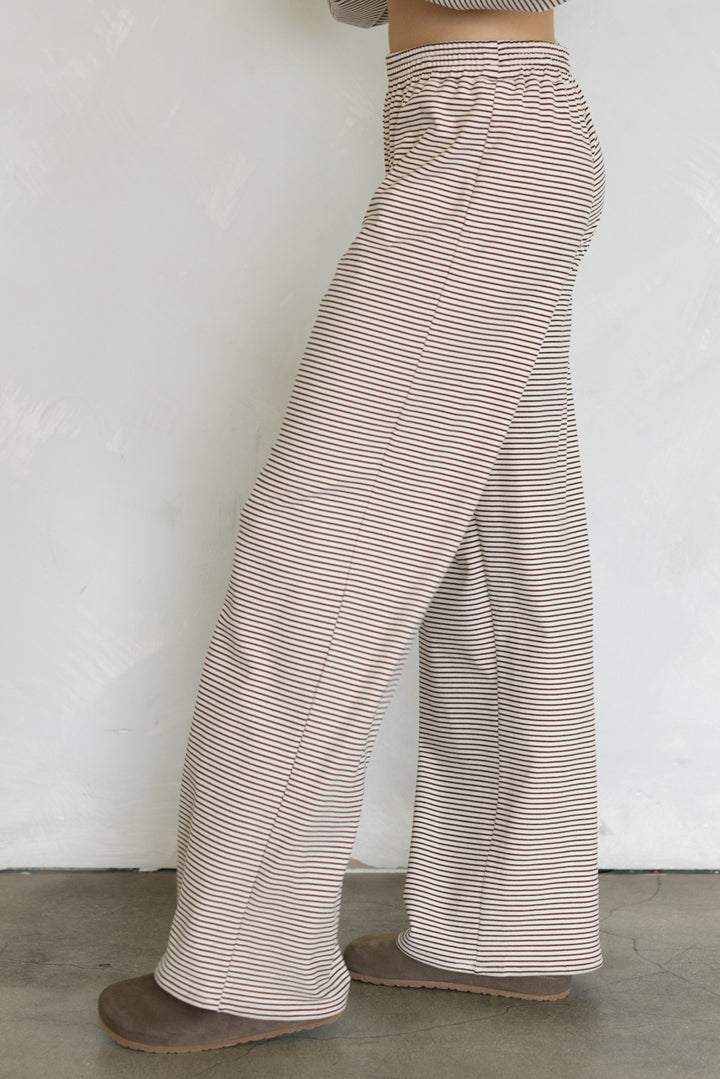 A person wearing loose, wide-leg pants with thin horizontal beige and brown stripes, shown from the waist down against a plain light wall.