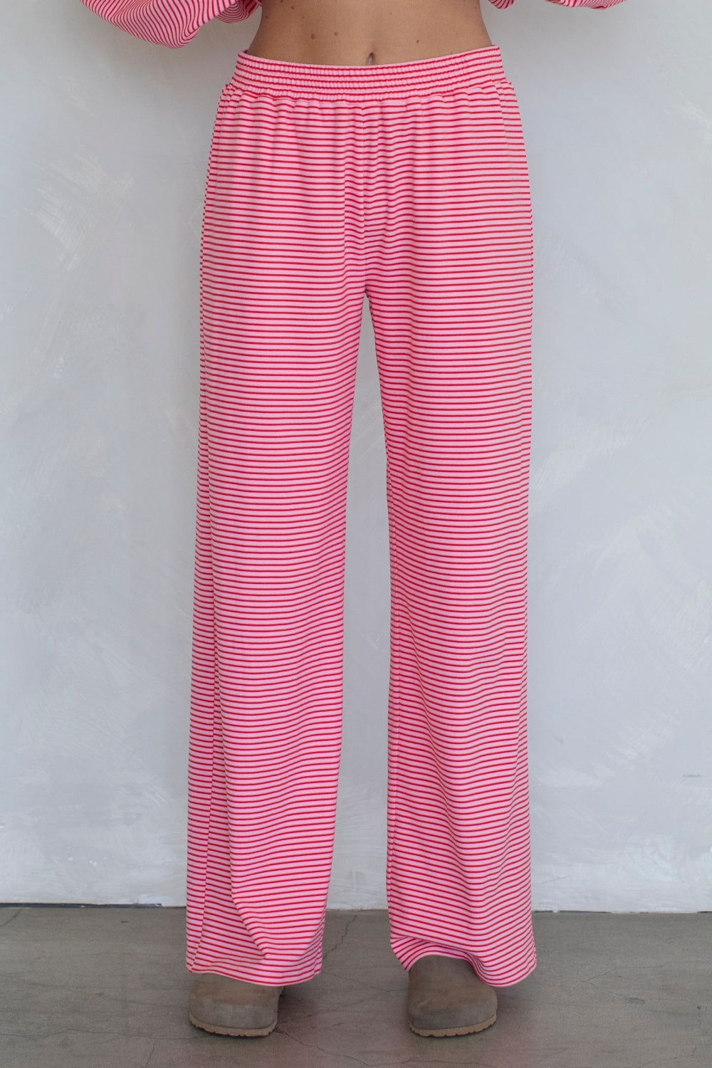 Pink and white striped wide-leg lounge pants shown on a model from the waist down, paired with gray slip-on shoes against a light gray wall.