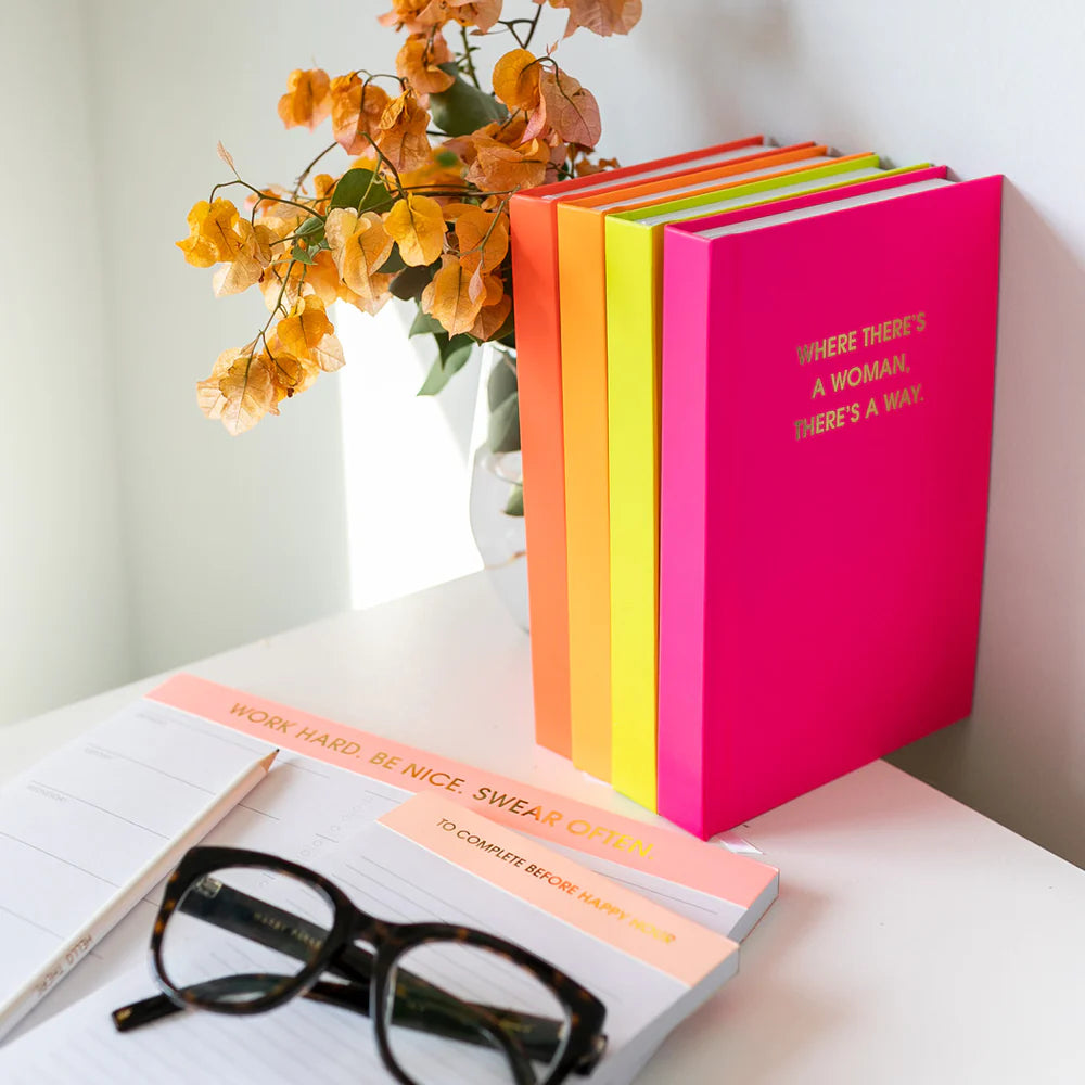 A set of brightly colored notebooks or journals standing upright on a white table, with one pink cover showing gold text. An open notebook with peach sticky notes, a pencil, and black-rimmed eyeglasses are also visible.
