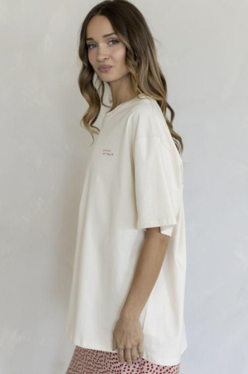 A woman wearing an oversized off-white t-shirt with small pink text on the left chest, paired with red and white patterned shorts, standing against a light gray textured wall.