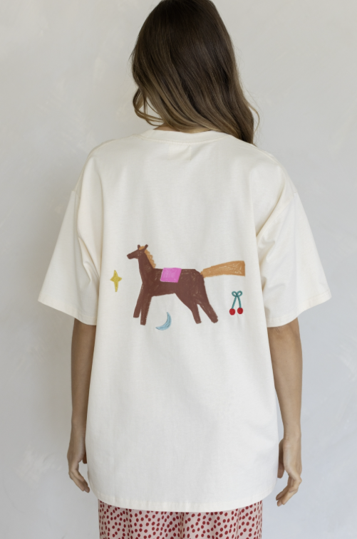 A woman viewed from behind wearing an oversized off-white t-shirt with a whimsical graphic of a brown horse with a pink blanket, a yellow star, and red cherries on the back. She is also wearing red and white patterned pants against a neutral light gray background.