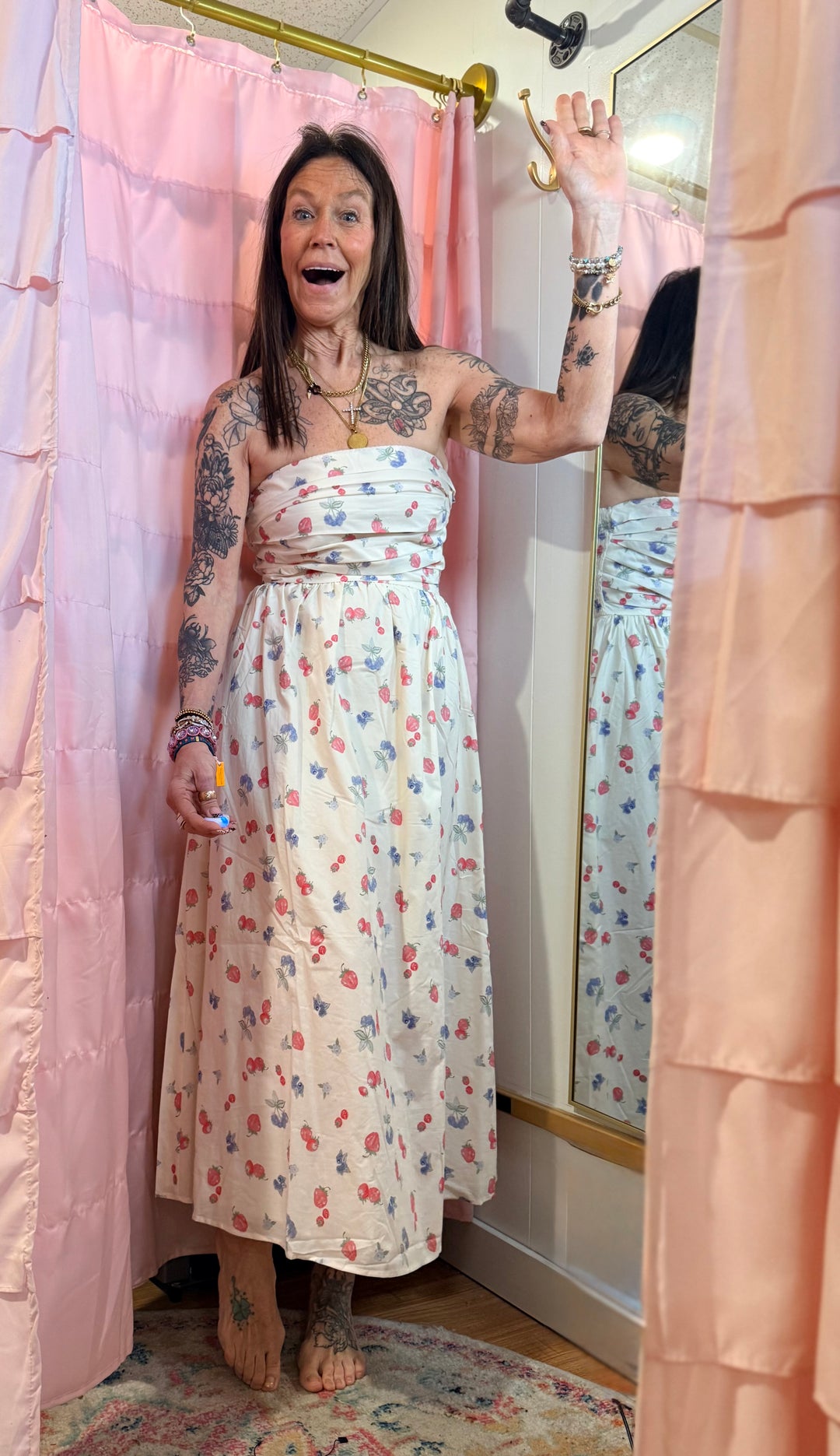 Woman in a floral dress standing in front of a mirror with pink curtains.