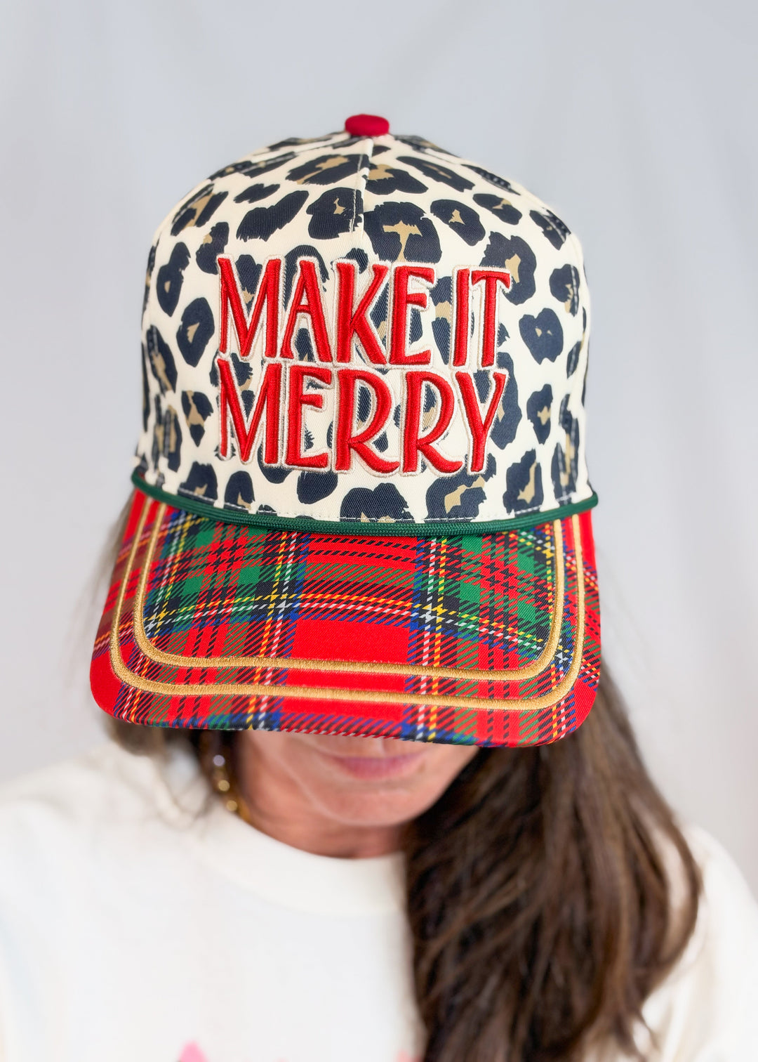 Woman wearing leopard print trucker hat with plaid bill and 'MAKE IT MERRY' embroidered on front on plain background