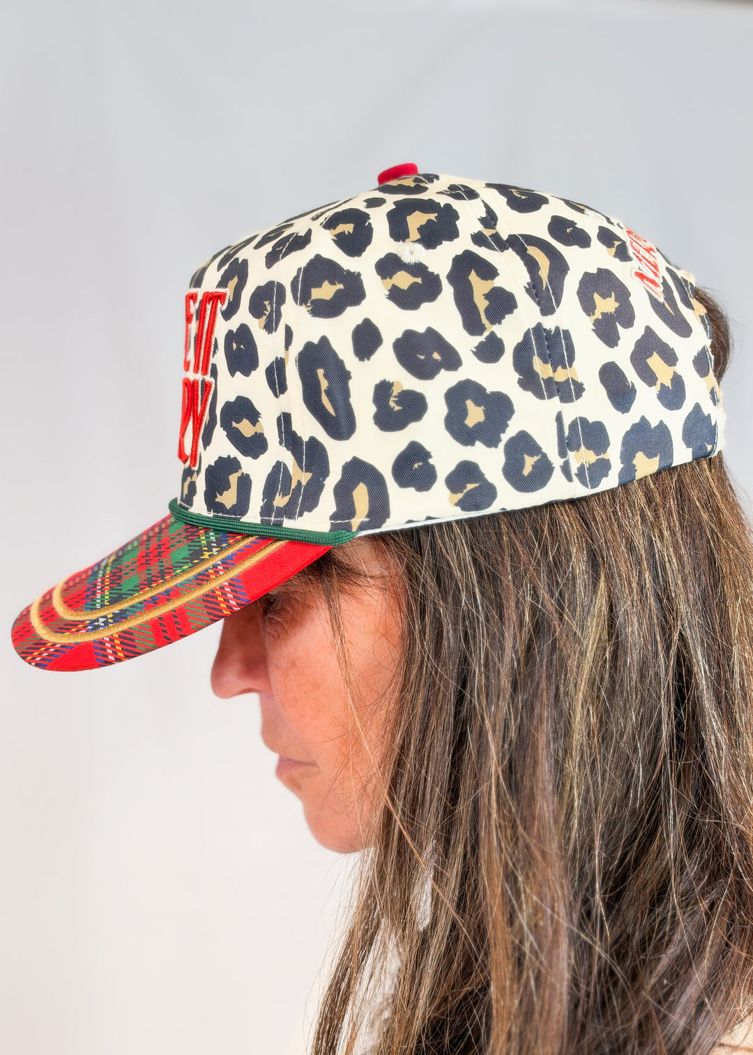 Woman shown from side view wearing leopard print trucker hat with plaid bill on plain background