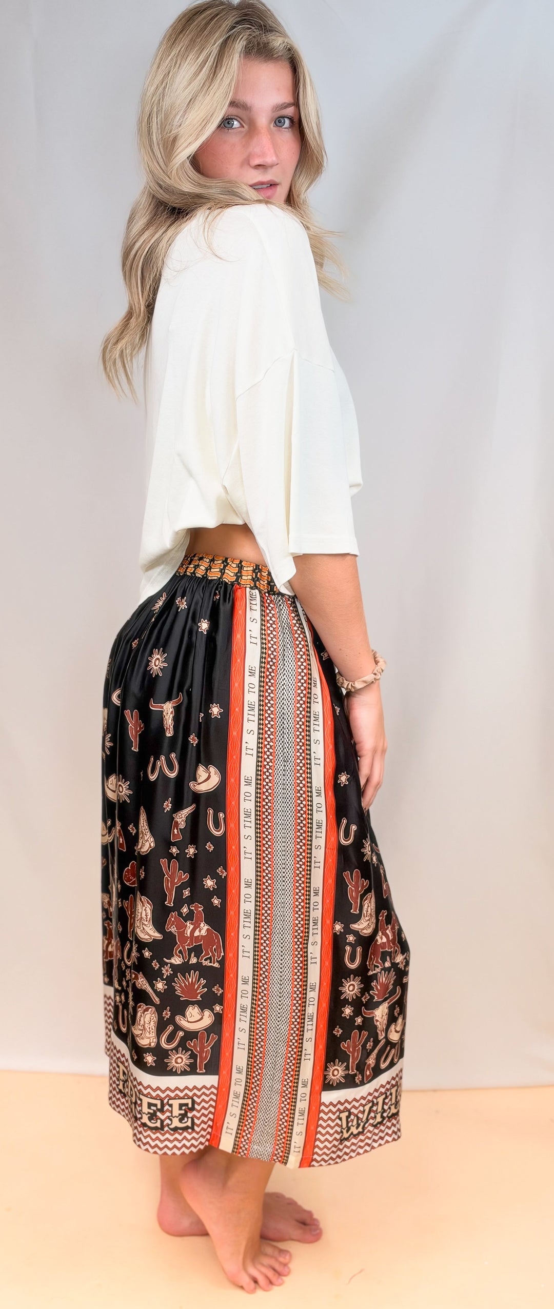 Woman shown from side view wearing white tee and rodeo motif scarf skirt on plain background