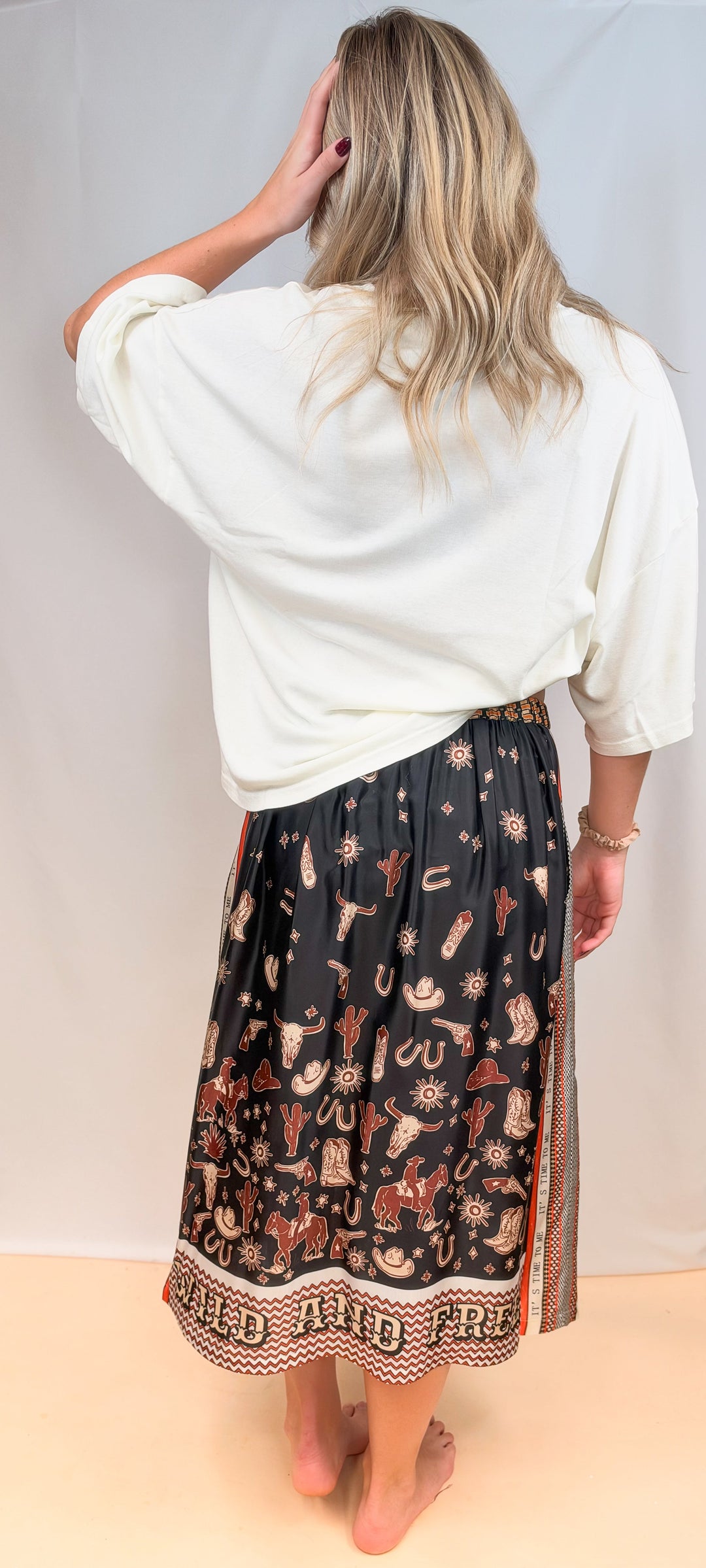 Woman shown from back view wearing white tee and rodeo motif scarf skirt on plain background