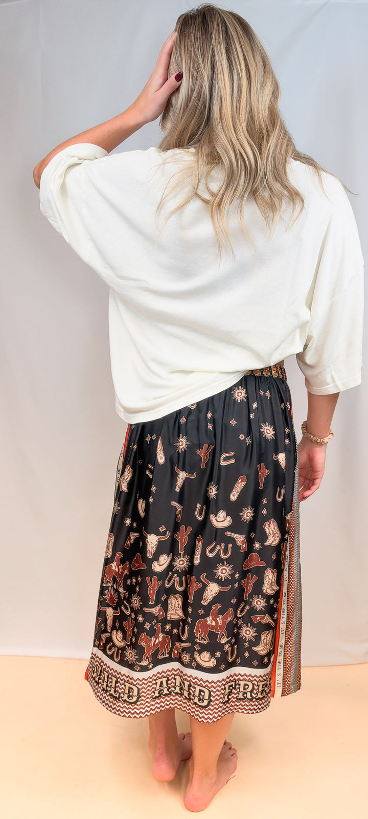 Woman shown from back view wearing white tee and rodeo motif scarf skirt on plain background