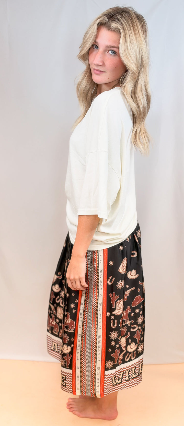 Woman shown from side view wearing white tee and rodeo motif scarf skirt on plain background