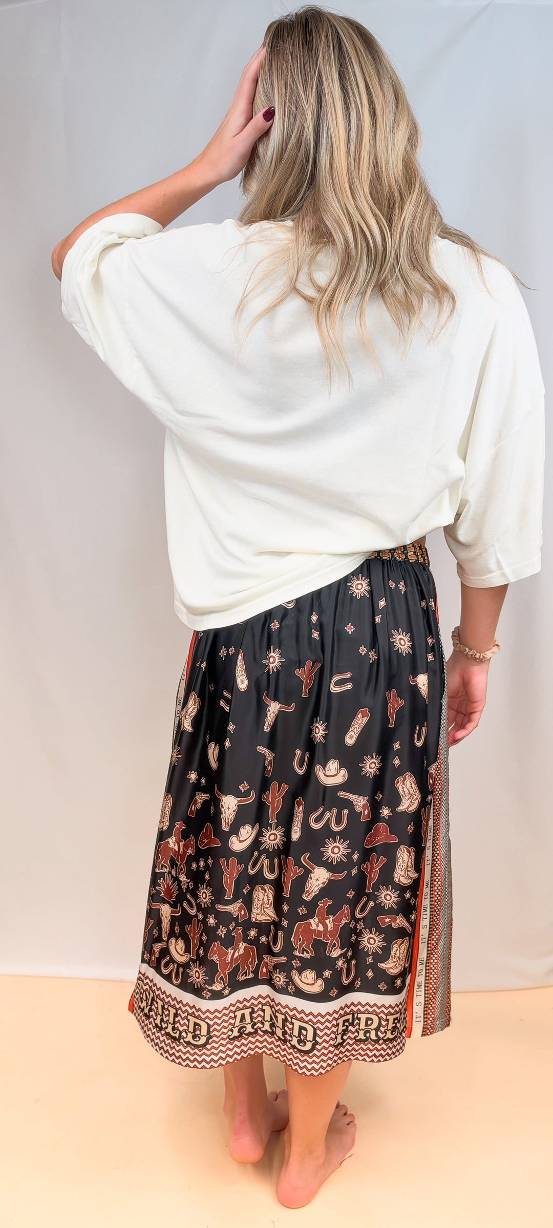 Woman shown from back view wearing white tee and rodeo motif scarf skirt on plain background