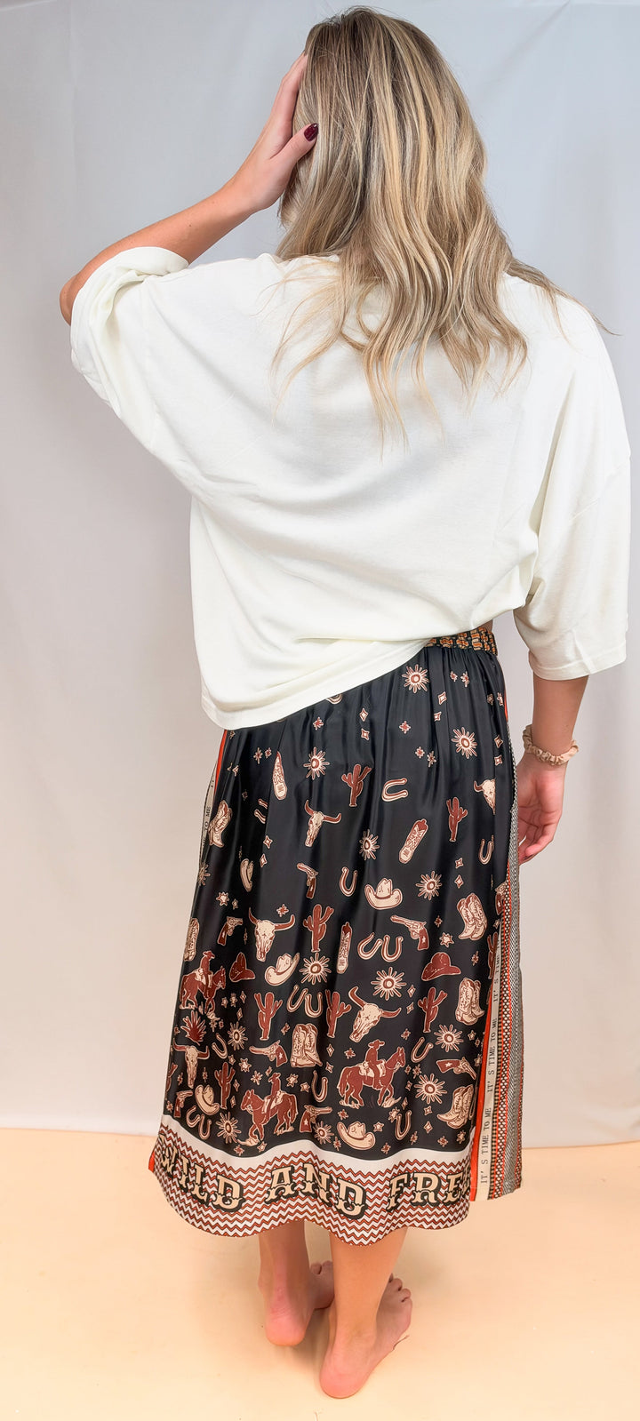 Woman shown from back view wearing white tee and rodeo motif scarf skirt on plain background