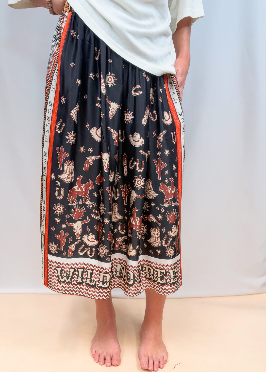 Woman shown from waist down wearing white tee and rodeo motif scarf skirt on plain background