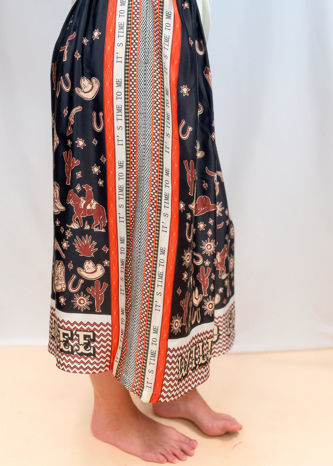 Woman shown from side view wearing white tee and rodeo motif scarf skirt on plain background