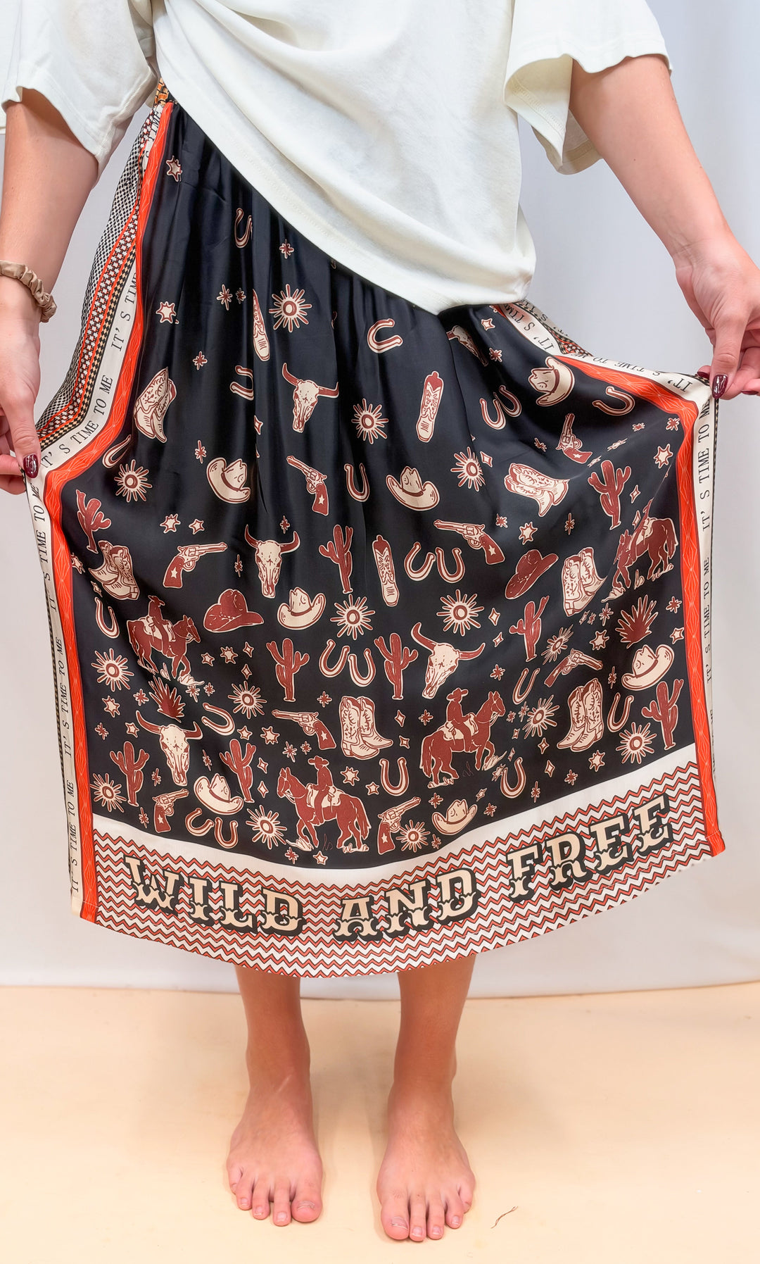 Woman shown from waist down wearing white tee and rodeo motif scarf skirt on plain background