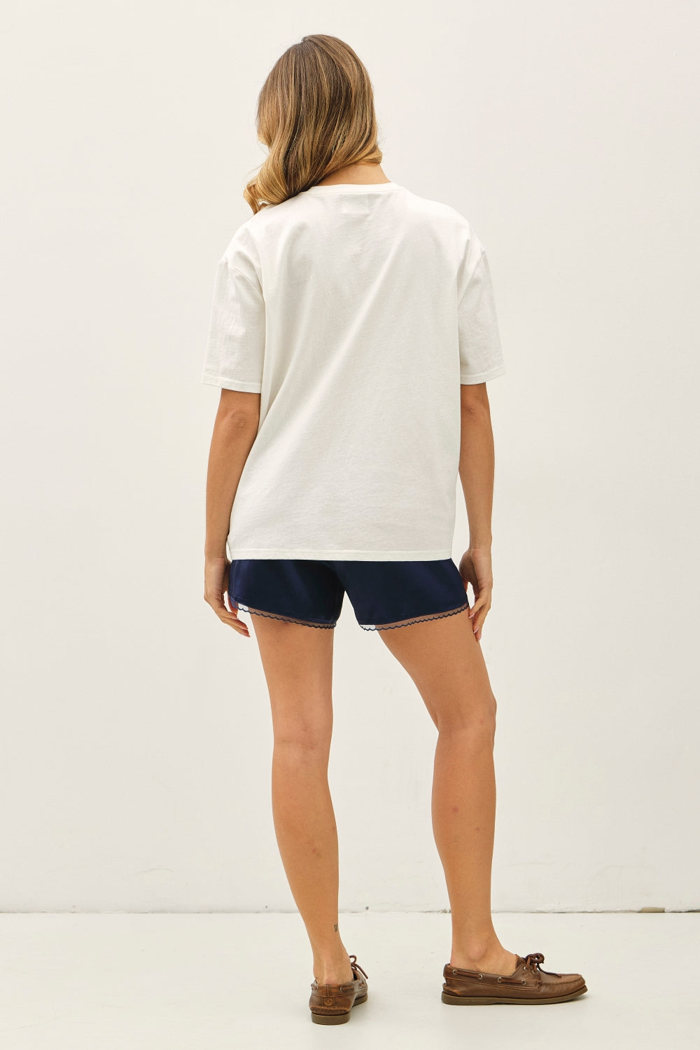 A woman shown from the back wearing a white short-sleeve t-shirt, navy blue shorts, and brown boat shoes against a plain white studio background.